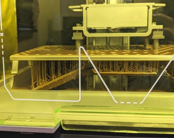 Può includere: Primo piano di una stampante 3D con un oggetto di colore dorato in fase di stampa. L'oggetto è un design complesso con molti dettagli intricati. La stampante è racchiusa in una custodia di plastica trasparente.