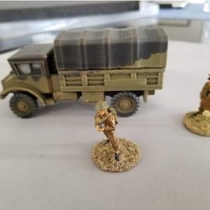 Puede incluir: Un camión militar en miniatura con una lona de lona que cubre la cama. El camión está pintado con un patrón de camuflaje y tiene dos figuras de pie cerca. Las figuras están pintadas con uniformes militares y sostienen rifles.