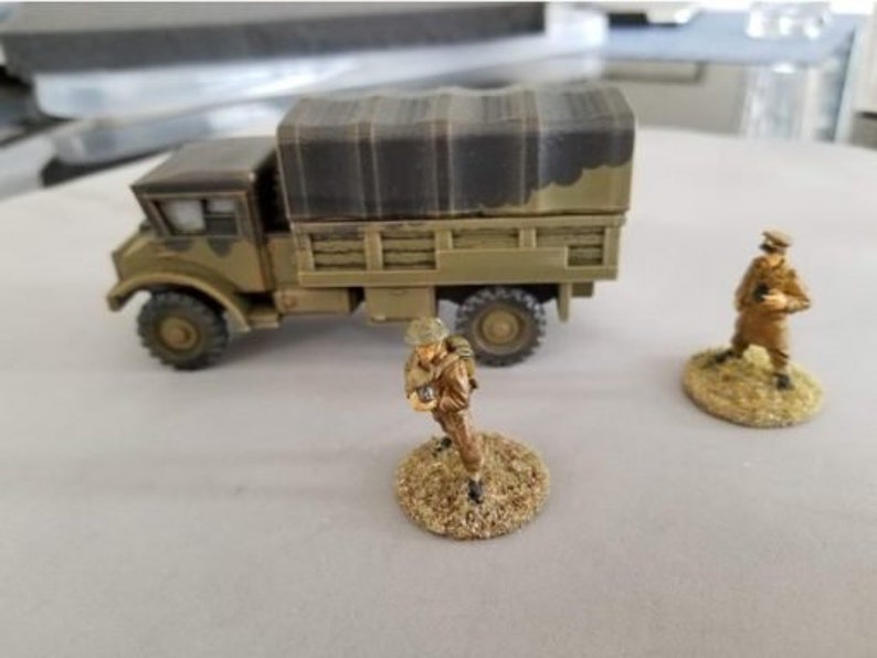 May include: A miniature military truck with a canvas tarp covering the bed. The truck is painted in a camouflage pattern and has two figures standing nearby. The figures are painted in military uniforms and are holding rifles.