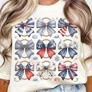 May include: Cream-colored t-shirt featuring a nautical bow design. The bows are in red, white, and blue, with rope accents, anchors, and seashells. The shirt has a relaxed fit.