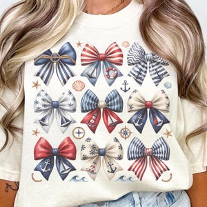 May include: Cream-colored t-shirt featuring a nautical bow design. The bows are in shades of red, white, and blue, with anchors, seashells, and nautical elements. The shirt has short sleeves and a relaxed fit.
