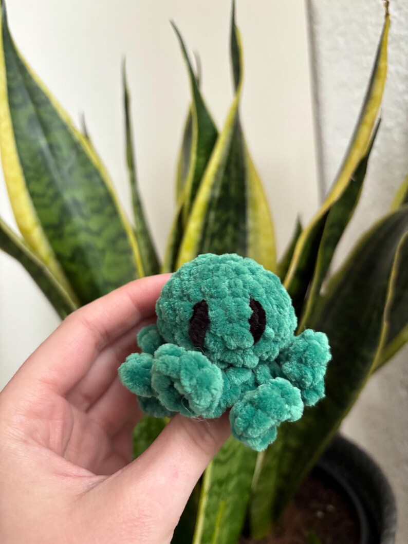 May include: A small green crocheted octopus with black eyes. The octopus is being held in a hand.