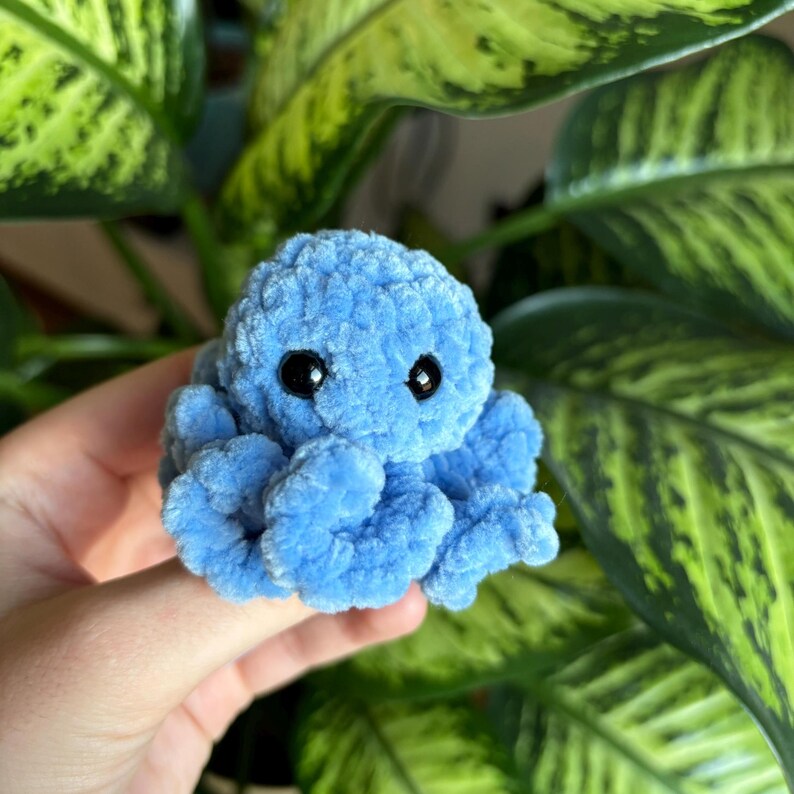 May include: A small blue crocheted octopus with black eyes. The octopus is sitting on a hand.