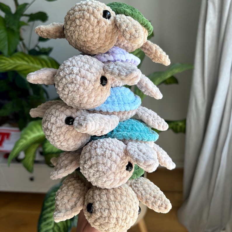 May include: Five crocheted turtle plush toys stacked on top of each other. Each turtle has a beige body, black eyes, and a shell in different colours: green, lavender, blue, and teal. The toys are arranged vertically, creating a whimsical display.
