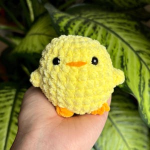 Könnte beinhalten: Ein kleines gelbes Häkeltier in Form eines Kükens mit schwarzen Augen und einem orangefarbenen Schnabel. Das Küken sitzt in einer Hand.
