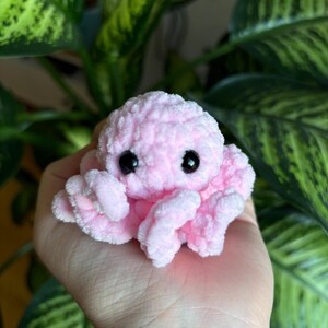 May include: A small pink crocheted octopus with black eyes. The octopus is sitting on a person's hand.