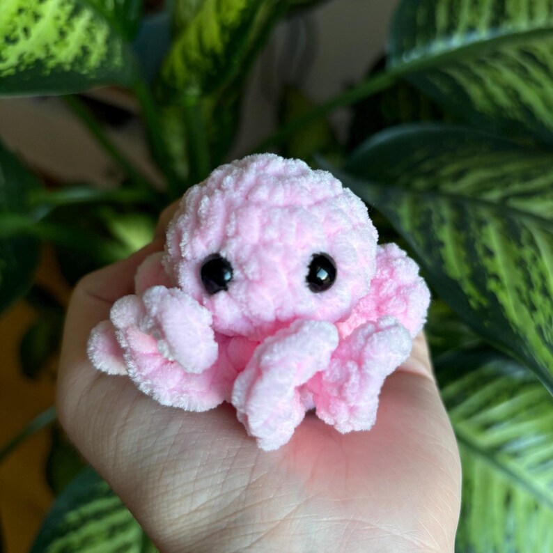 May include: A small pink crocheted octopus with black eyes. The octopus is sitting on a person's hand.