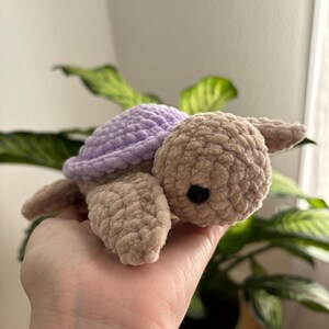 May include: A handmade crochet turtle in soft, textured yarn. The turtle has a light purple shell, a tan body, and small black eyes. The plush toy is designed with a cute, friendly appearance.