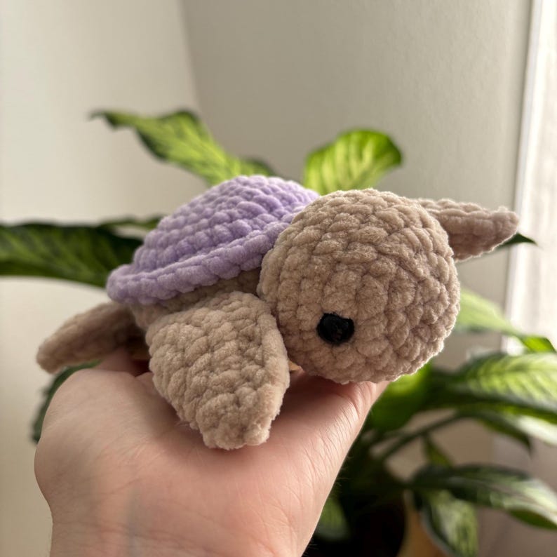 May include: A handmade crochet turtle in soft, textured yarn. The turtle has a light purple shell, a tan body, and small black eyes. The plush toy is designed with a cute, friendly appearance.