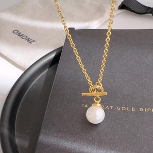May include: A gold chain necklace with a toggle clasp and a single white pearl pendant. The necklace is displayed on a black surface with a white box that says "OMONZ" and "14 karat gold dipped".