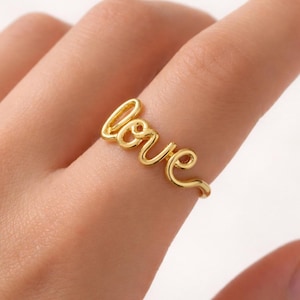 May include: A gold-colored ring spelling out the word "love" in a cursive script. The ring is worn on a finger, showcasing its delicate design and warm hue. The ring's elegant simplicity makes it a charming accessory.