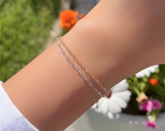 May include: A delicate two-tone bracelet featuring one thin gold rope chain and one thin silver rope chain worn together. The chains have a twisted, textured design that catches the light, creating a layered appearance on the wrist.