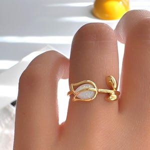 May include: A gold-colored ring in the shape of a tulip, with a white opal stone in the center. The ring is open and adjustable, with leaf details on the band. The ring is worn on a finger.