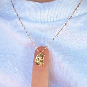 May include: A gold-colored pendant necklace with a small, abstract design. The pendant is suspended from a delicate chain. The necklace is displayed on a finger, against a white t-shirt with a pink and blue graphic.