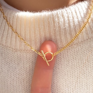 May include: A gold-colored necklace with a toggle clasp. The necklace features two different chain styles: a delicate cable chain and a rectangular link chain. The clasp is a small bar that fits through a circular loop.