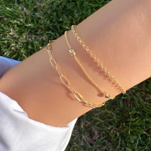 May include: Three gold bracelets of varying designs are displayed on an arm against a backdrop of green grass. The bracelets include a chain link, a delicate chain with a small rectangular charm, and a beaded chain. The arm is wearing a white shirt.