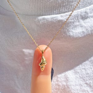May include: A delicate gold necklace featuring a small seashell pendant. The pendant is intricately detailed and hangs from a fine gold chain. The necklace is displayed against a white t-shirt.