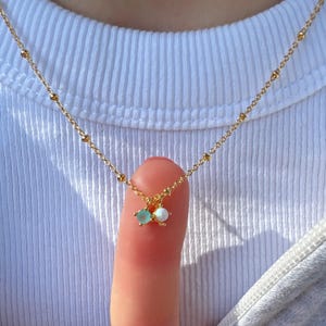 May include: A delicate gold necklace with a beaded chain and two small charms. One charm is a light blue gemstone, and the other is a white opal. The necklace is displayed against a white background.