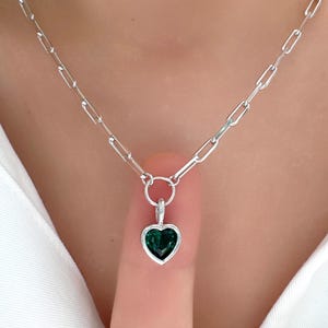 May include: A silver necklace with a heart-shaped pendant. The pendant is a dark green color and is set in a silver frame. The chain is a link style. The necklace is displayed against a white shirt.