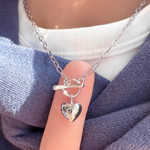 May include: A silver-colored necklace with a heart-shaped pendant. The necklace features a toggle clasp and a delicate chain. The heart pendant is smooth and reflective, hanging from the chain. The necklace is shown against a blue background.