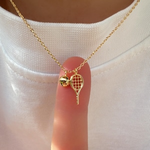 May include: A gold chain necklace with a tennis racket and ball charm. The charm is a small, detailed representation of a tennis racket and ball.
