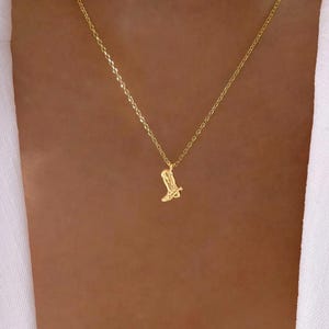 May include: A delicate gold-colored necklace featuring a small pendant shaped like a cowboy boot. The pendant hangs from a fine chain, creating a simple yet stylish accessory. The necklace is shown against a neutral background.