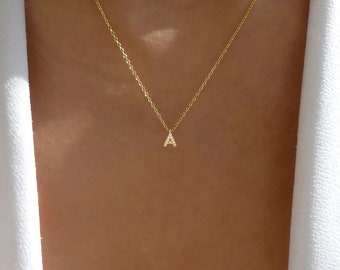 May include: A delicate gold chain necklace featuring a small, dainty pendant in the shape of the capital letter A. The letter pendant is embellished with tiny, clear, sparkling stones set along its surface, creating a subtle and refined appearance.