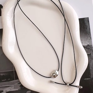 May include: A minimalist necklace featuring a black cord and a silver-toned, irregularly shaped pendant. The necklace also includes a small, cylindrical silver bar. The jewelry is displayed on a white, organically shaped dish.