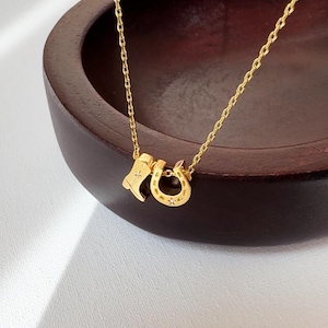 May include: A gold-colored necklace featuring a small cowboy boot charm and a horseshoe charm. The necklace is displayed against a dark wooden background and a white surface.