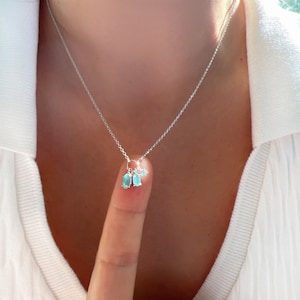 May include: A silver chain necklace with a small pendant featuring three blue gemstones. The pendant is held by a finger.
