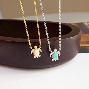 May include: Two delicate necklaces with turtle pendants. One necklace has a gold chain and a white opal turtle pendant. The other necklace has a silver chain and a blue opal turtle pendant.