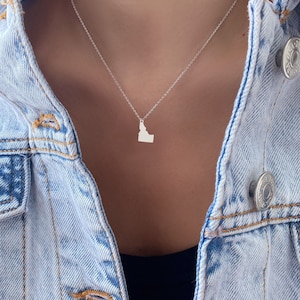 May include: A silver necklace with a pendant shaped like the state of Idaho. The necklace is worn by a person wearing a denim jacket.