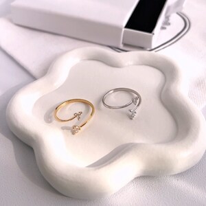 May include: Two open-ended rings with a leaf-like design, one gold-toned and the other silver-toned, each adorned with small, clear gemstones. The rings rest on a white, organically shaped tray, with a white box in the background.