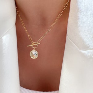 May include: Gold-toned necklace featuring a rectangular pendant with a clear, faceted stone. The necklace has a toggle clasp and a delicate chain. The pendant is set in a gold frame, creating a classic and elegant look.