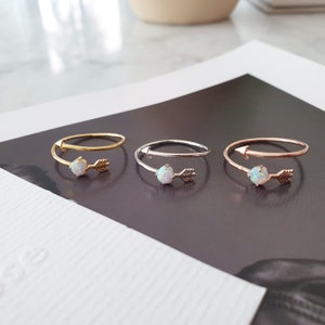 May include: Three delicate rings with a white opal stone and a gold arrow design. The rings are shown on a white surface.