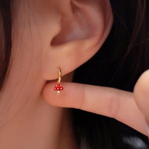 May include: Gold hoop earrings with a small red and white mushroom charm hanging from the hoop.