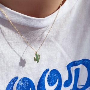 May include: A gold chain necklace with a green and gold cactus pendant. The necklace is worn by a person wearing a white t-shirt with blue text.