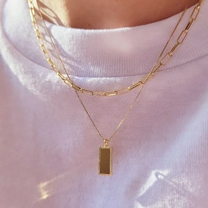 May include: A gold chain necklace with a rectangular pendant. The chain is layered with a thicker chain.