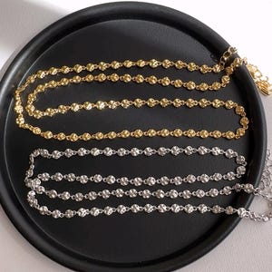 Puede incluir: Dos cadenas delicadas, una dorada y otra plateada, con pequeños eslabones redondos texturizados. Las cadenas se muestran en un plato negro.