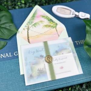 May include: A white envelope with a green ribbon and a wax seal. The envelope is decorated with a watercolor painting of palm trees and a sunset. The text "Key West" is written on the envelope. The envelope is sitting on a blue book with the text "National Geographic" visible.