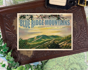 Può includere: Una carta rettangolare con un'illustrazione paesaggistica delle Blue Ridge Mountains al tramonto. Il testo recita Meet us in the BLUE RIDGE MOUNTAINS e KRISTIN & JACKSON - 09.17.2027. La carta è posizionata su una superficie in pelle marrone.
