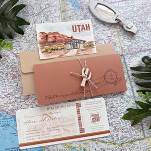 May include: A wedding invitation suite featuring a boarding pass, a postcard with "UTAH" text, and a terracotta envelope tied with twine and a wooden cactus charm. The invitation suite is arranged on a map.