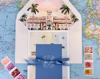 Puede incluir: Un conjunto de invitaciones con una ilustración en acuarela de un edificio y palmeras, presentado dentro de un sobre blanco. El conjunto incluye una tarjeta azul con un monograma, atada con una cinta azul y sellos florales. El fondo es un mapa del mundo.