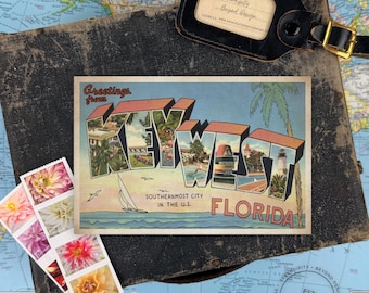 Può includere: Una cartolina in stile vintage con il testo Greetings from KEY WEST FLORIDA SOUTHERNEST CITY IN THE U.S. e illustrazioni di una barca a vela, palme e un faro, accompagnata da un'etichetta per bagagli nera e una striscia di francobolli floreali.