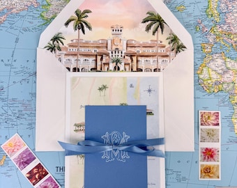 Può includere: Un set di cancelleria composto da una busta con un'illustrazione ad acquerello di un grande edificio e palme, una mappa con l'etichetta Treasure Coast e un faro, e un cartoncino blu con un monogramma bianco legato con un nastro di raso blu.