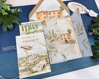 Può includere: Un set di partecipazioni di nozze con illustrazioni ad acquerello di una villa e un paesaggio italiano. Include una velina con motivi di rami d'ulivo, un cartoncino con il testo Carla & Fredrik e un piccolo ciondolo in legno.