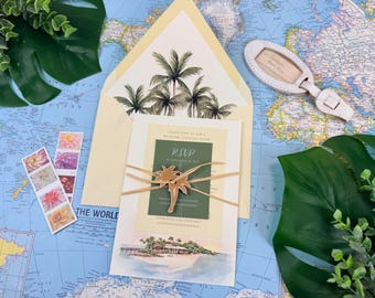 Puede incluir: Un conjunto de invitaciones con temática tropical. La invitación tiene una ilustración en acuarela de una escena de playa y está atada con hilo. El sobre tiene un diseño de palmera. También se incluyen sellos de flores y una etiqueta de equipaje.