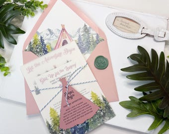 Può includere: Una busta rosa con un'illustrazione ad acquerello di montagne e un tepee sulla fodera, contenente biglietti d'invito legati con spago blu e bianco. È attaccata un'etichetta per lotteria in carta a forma di tepee e un sigillo in cera verde.