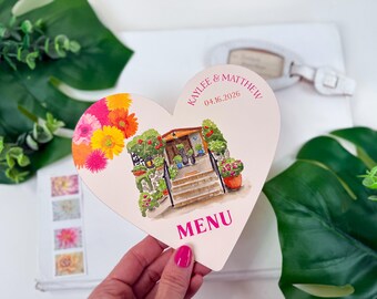 Puede incluir: Una tarjeta de menú en forma de corazón con una ilustración de acuarela de un edificio con escaleras, flores y la palabra "MENU". La tarjeta tiene los nombres "Kaylee & Matthew" y la fecha "04.16.2026" impresos. La tarjeta está decorada con flores coloridas.
