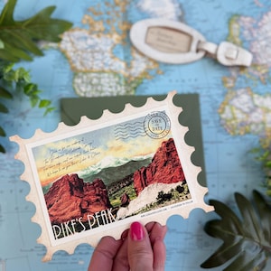 May include: A rectangular save the date card shaped like a postage stamp featuring a landscape illustration of red rock formations and a mountain peak labeled PIKE'S PEAK. The card includes handwritten-style text and a circular postmark stamp.